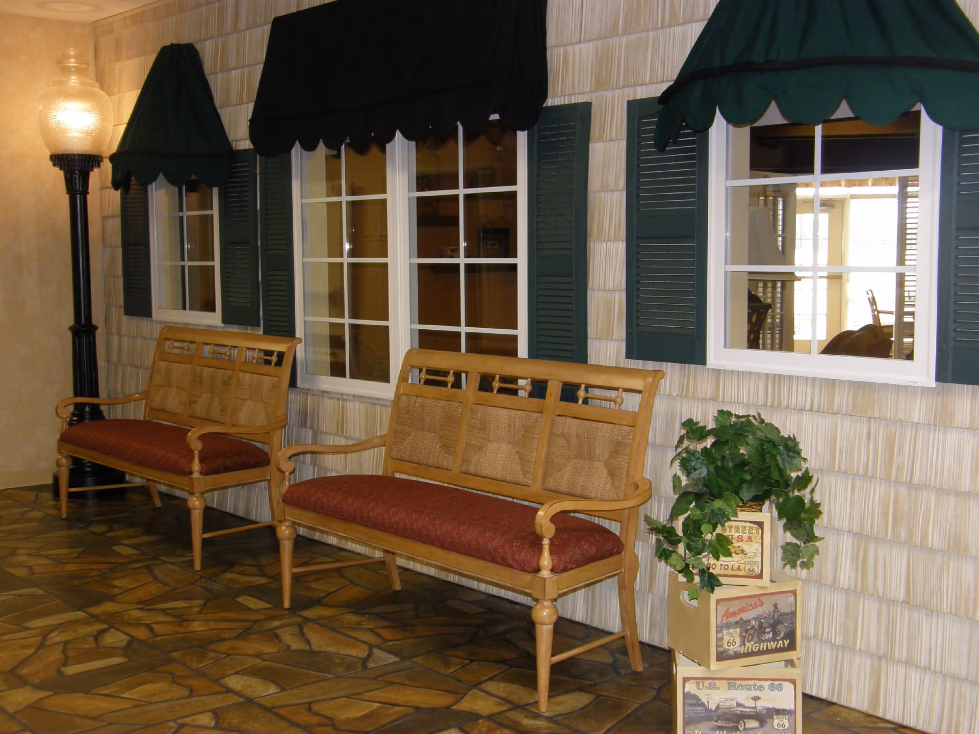 Indoor seating area with two wooden benches against a faux exterior wall with windows, green awnings, a floor lamp, and a potted plant.