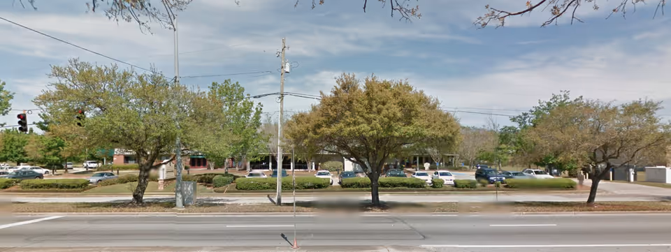 Wide street view of a facility's exterior grounds with trees, parked cars, and landscaping.