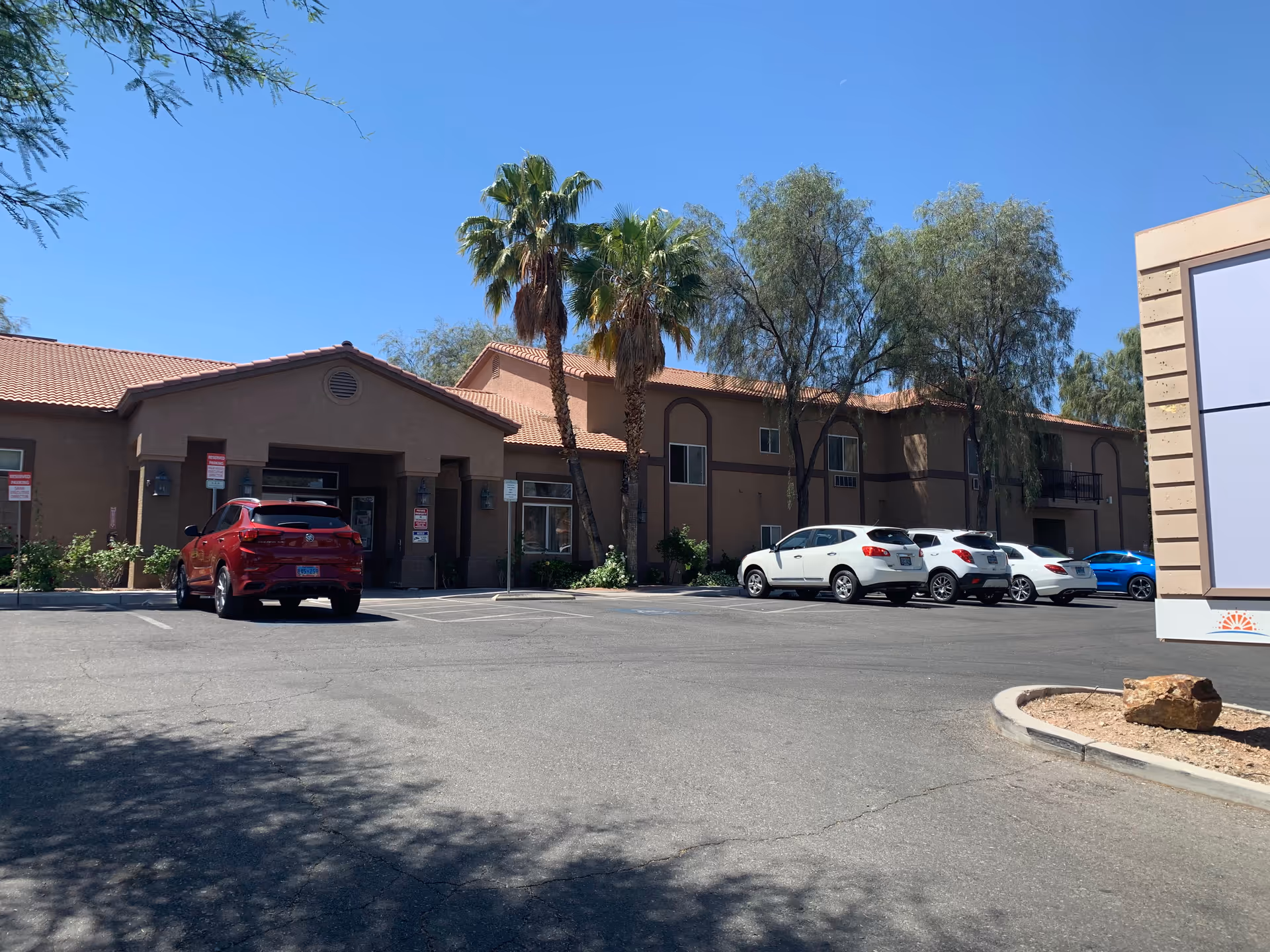Exterior view of Sana Assisted Living facility showing a two-story building with a tiled roof, palm trees, and several parked cars in the parking lot under a clear blue sky.