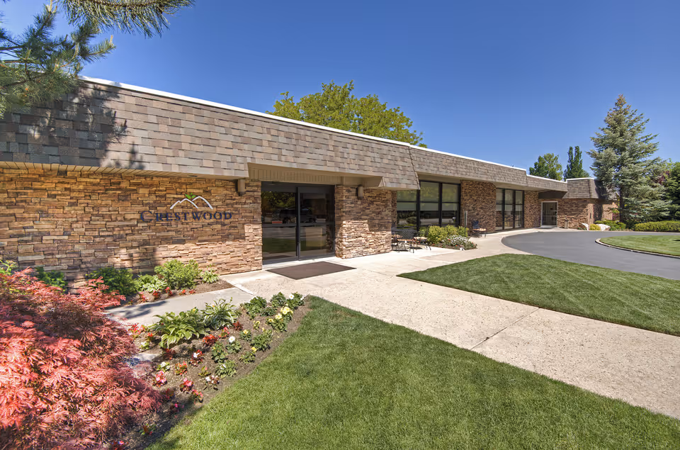 Exterior view of Crestwood Rehabilitation and Nursing facility showing a single-story building with stone facade, large windows, a glass entrance door, landscaped garden with flowers and shrubs, and a paved walkway and driveway under a clear blue sky.