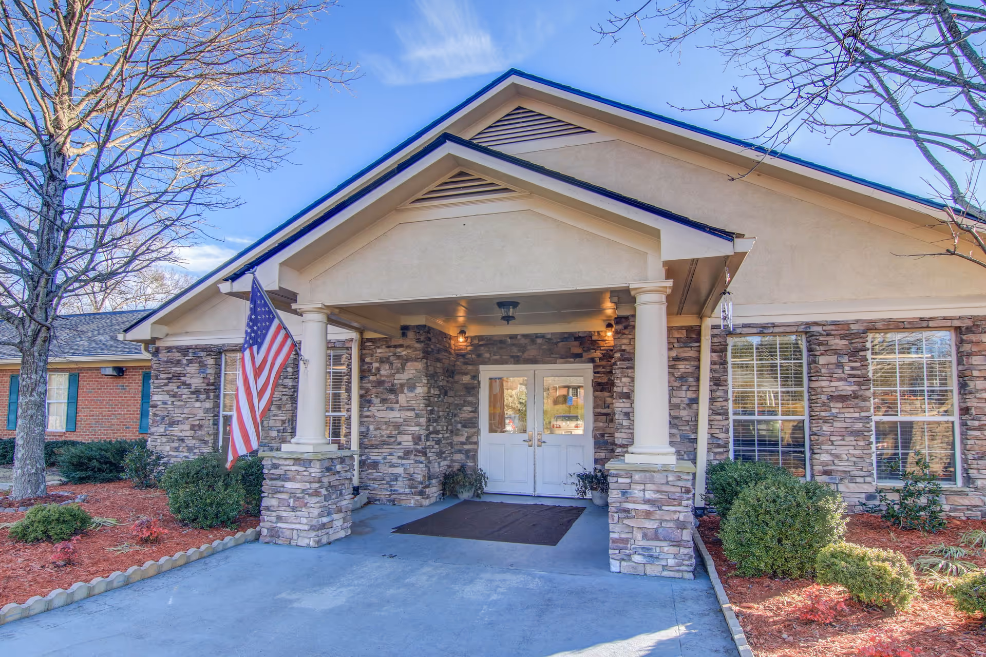 Front entrance of Manchester Court Assisted Living & Memory Care featuring a covered porch with stone pillars, an American flag mounted on the left pillar, double white doors, and landscaped bushes and trees around the building.