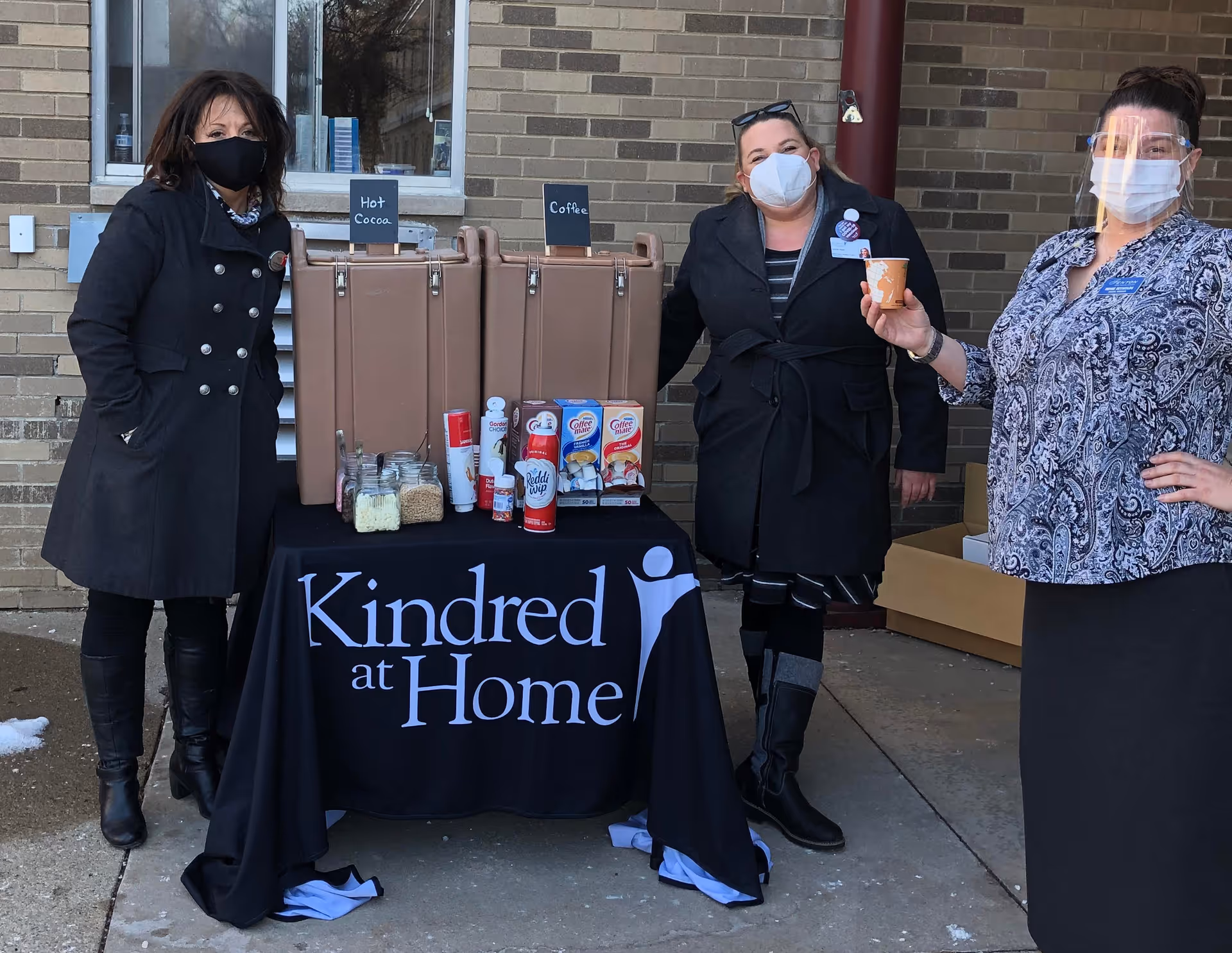 Three women wearing face masks and standing outside next to a table covered with a black cloth that reads 'Kindred at Home'. On the table are two large beverage dispensers labeled Hot Cocoa and Coffee, along with various coffee and hot cocoa supplies. The women are dressed warmly and one is holding a cup.