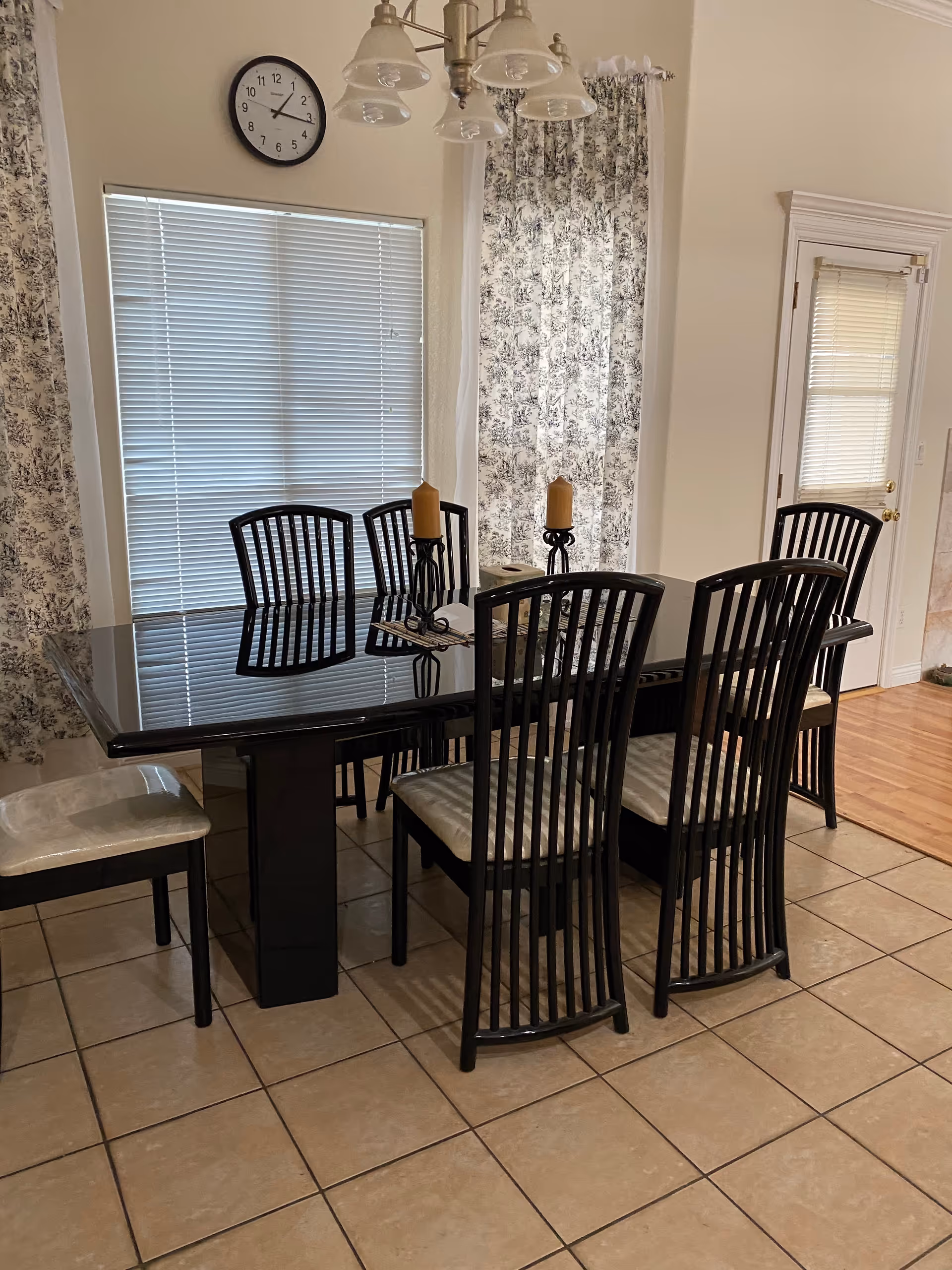 Glass-top dining table with six black chairs, candles, blinds, and floral curtains in a dining room.