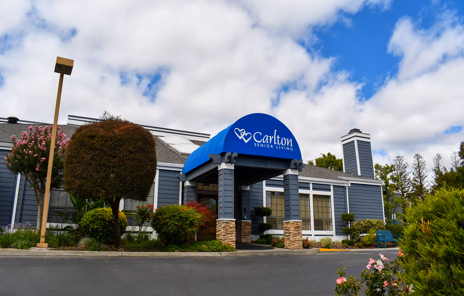 Exterior view of Carlton Senior Living Pleasant Hill - Martinez building with a blue awning displaying the facility's name. The building is gray with white trim and surrounded by landscaped bushes, trees, and flowers under a partly cloudy sky.