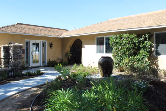 Single-story beige senior living facility with arched entrance and lush courtyard landscaping.