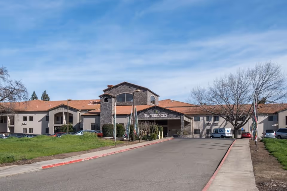 Front entrance and driveway of 'The Terraces' senior living building with a red-tiled roof, flags, and parked cars.