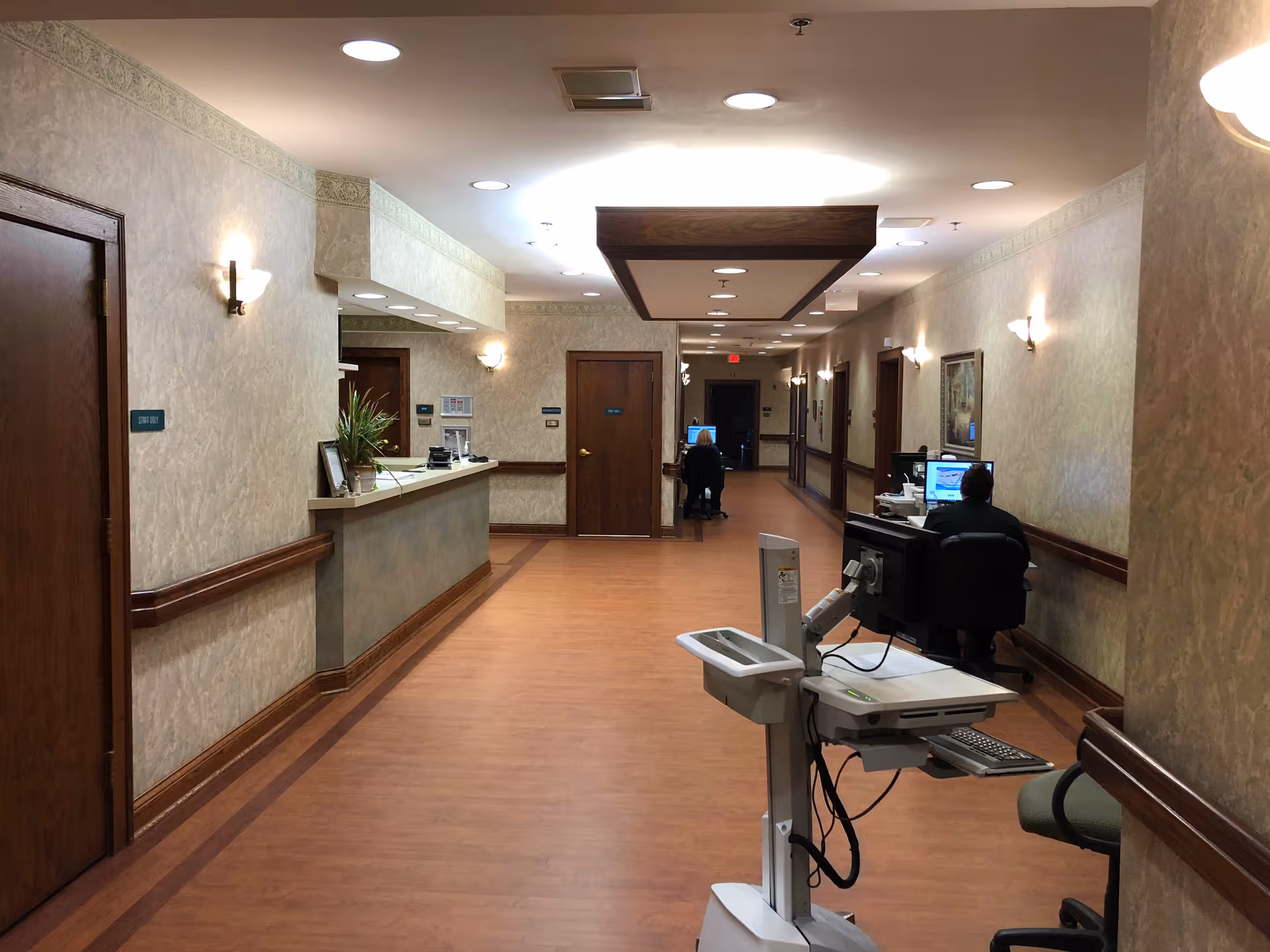 Long, well-lit hallway in a hospice care center with a reception desk and staff.
