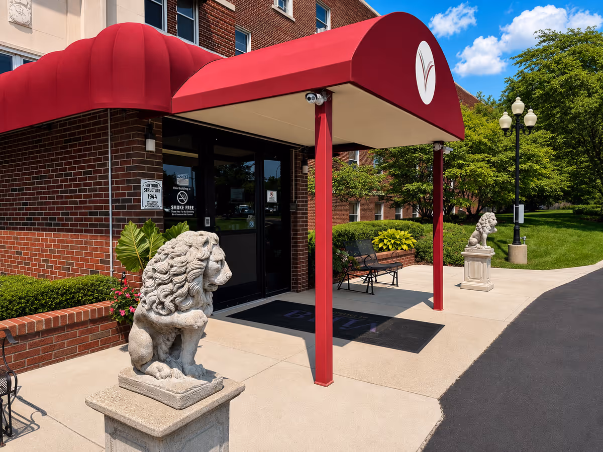 Brick senior living entrance with red canopy, lion statues, benches, and greenery.