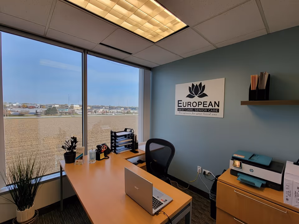 Office desk with laptop, printer, and a European Best Care sign by a large window.