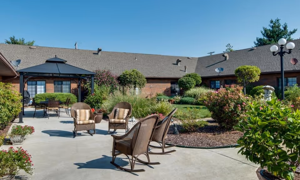 Sunlit courtyard with wicker seating and a gazebo surrounded by landscaped gardens in front of a one-story brick building.