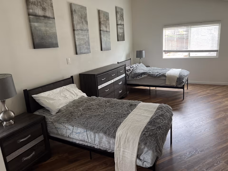 Two single beds with gray blankets and a dresser, in a bright bedroom.