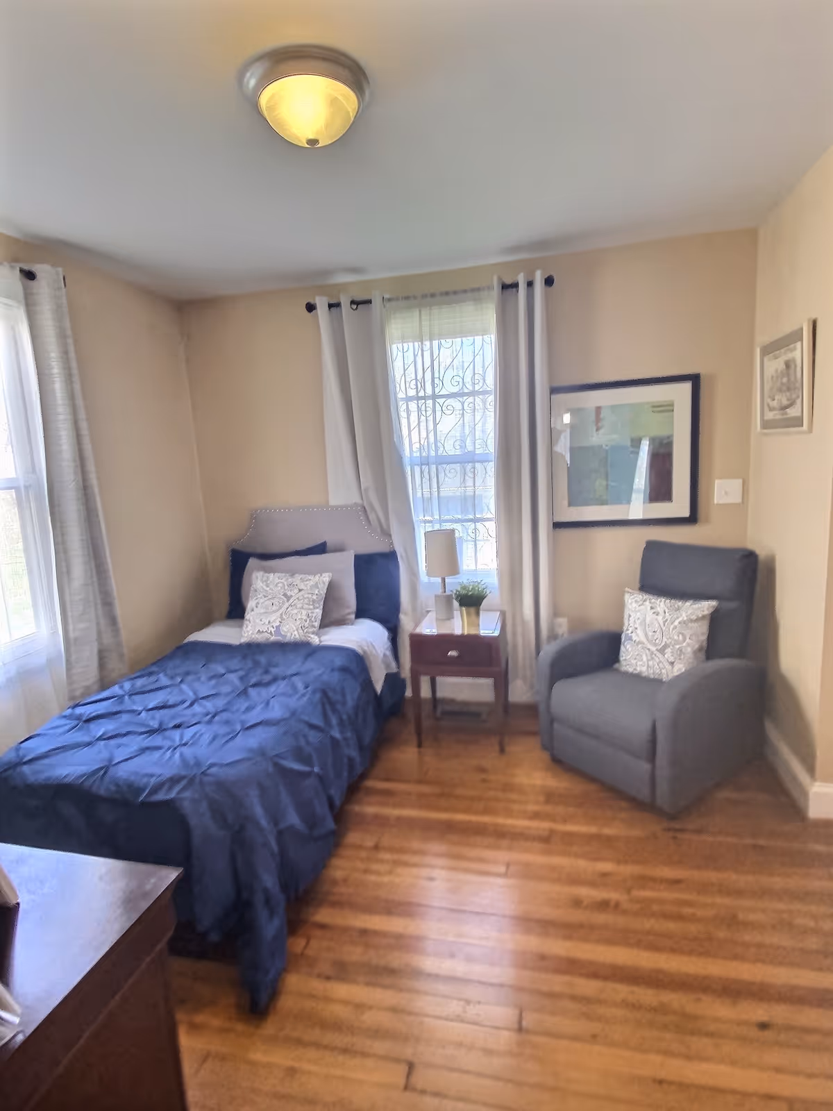 Cozy bedroom with blue quilted bed, nightstand, lamp, and gray armchair.