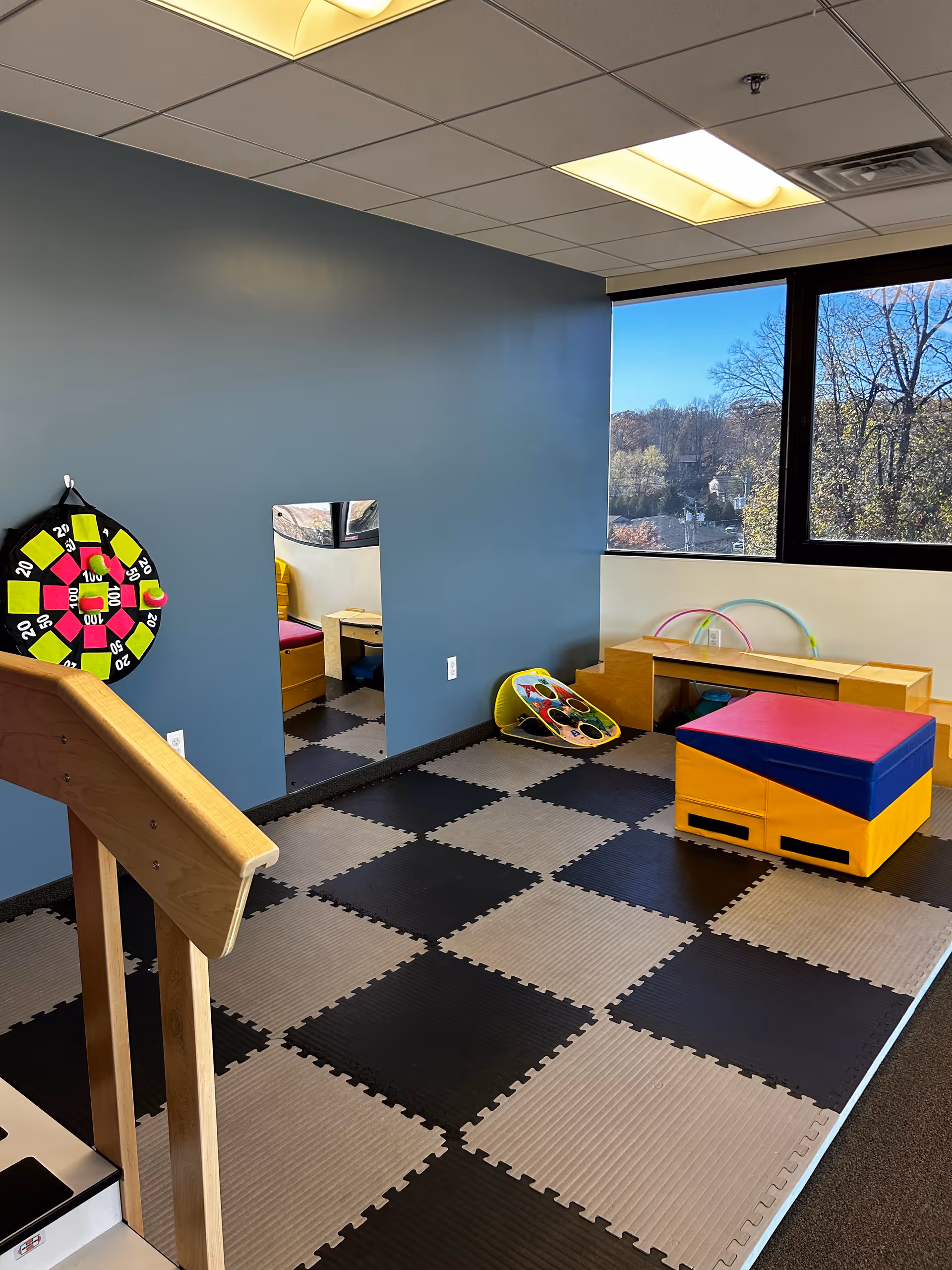 Interior sensory gym with blue wall, checkered foam flooring, window, and colorful play equipment.