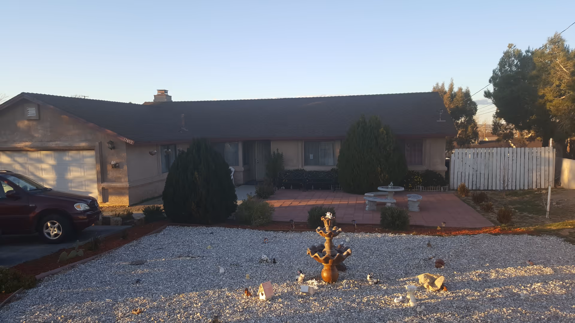 Single-story residential building with a dark roof, beige exterior walls, and a garage on the left side. In front of the building is a gravel yard with a decorative fountain in the center, surrounded by small plants and bushes. A maroon vehicle is parked in the driveway to the left. There are two large bushes near the entrance and a white wooden fence on the right side. The sky is clear and the scene is lit by late afternoon sunlight.