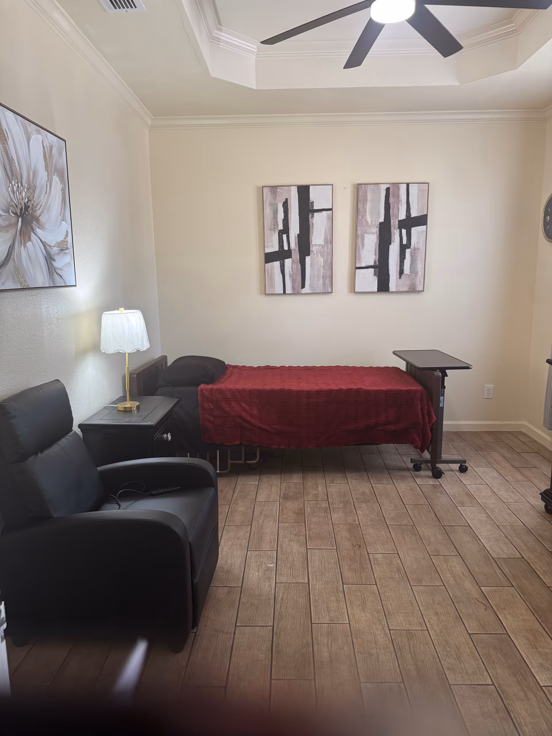 A compact senior-living bedroom with a bed, armchairs, lamp, and abstract art.