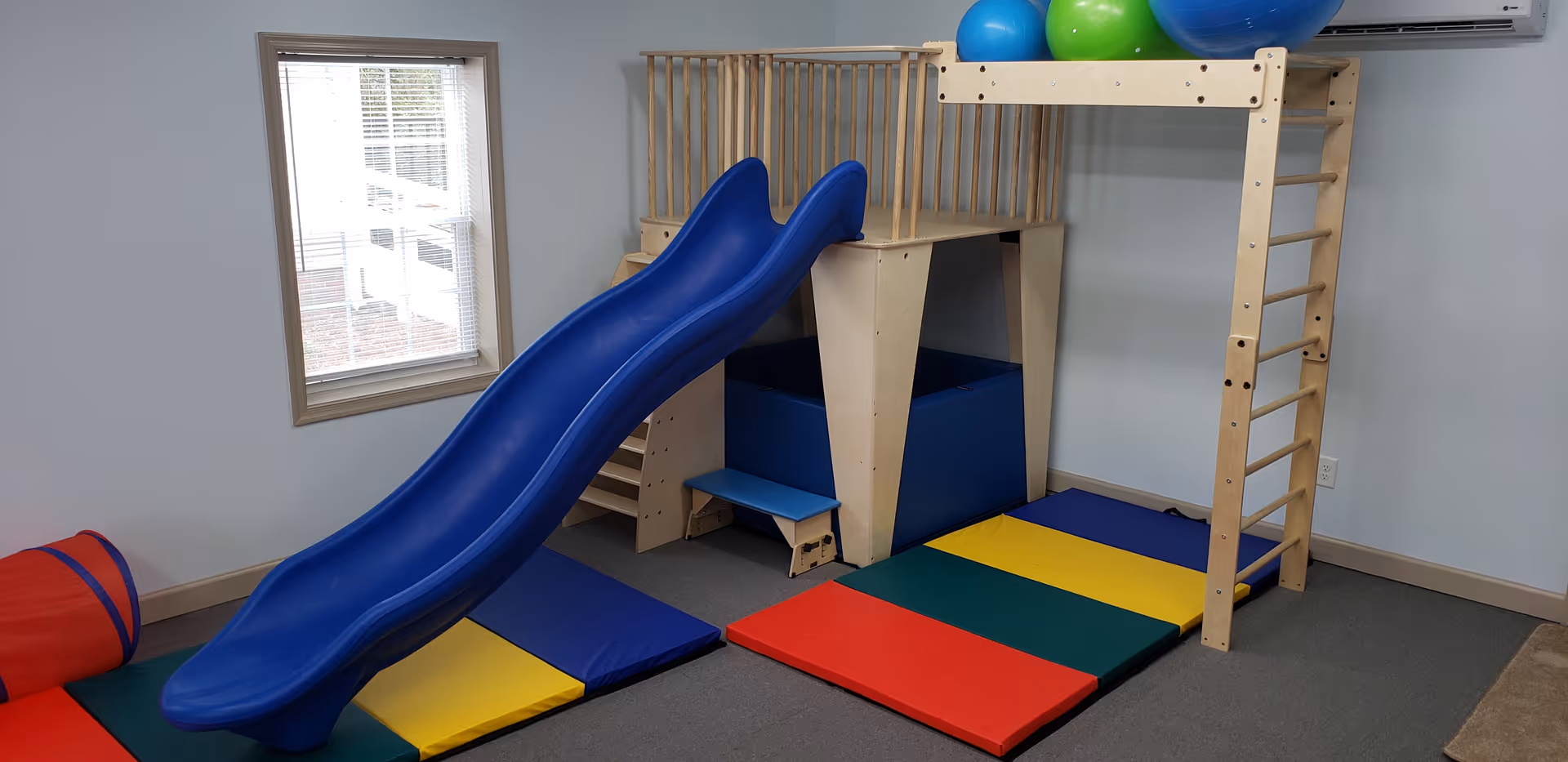 Indoor play area with a wooden climbing frame, blue slide, and colorful mats.