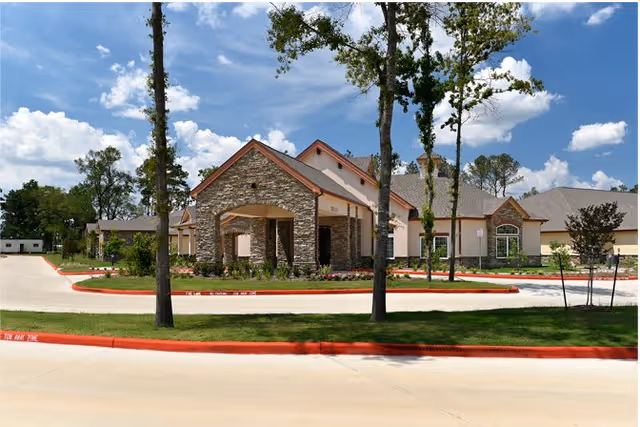 Exterior view of a senior living community building with stone and beige siding, surrounded by trees and landscaping under a blue sky with scattered clouds.