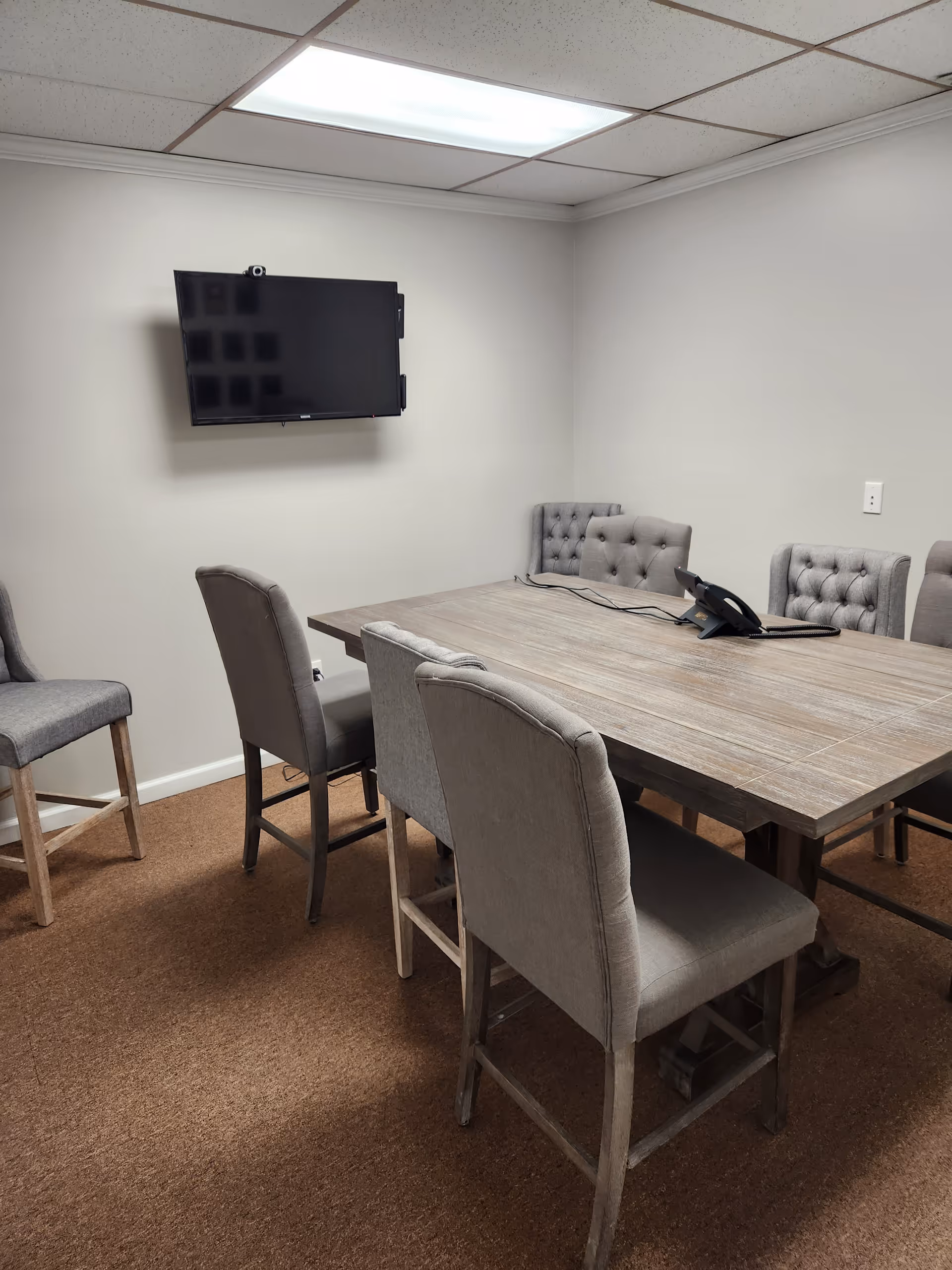A rectangular conference table with upholstered chairs and a wall-mounted TV in a plain room.
