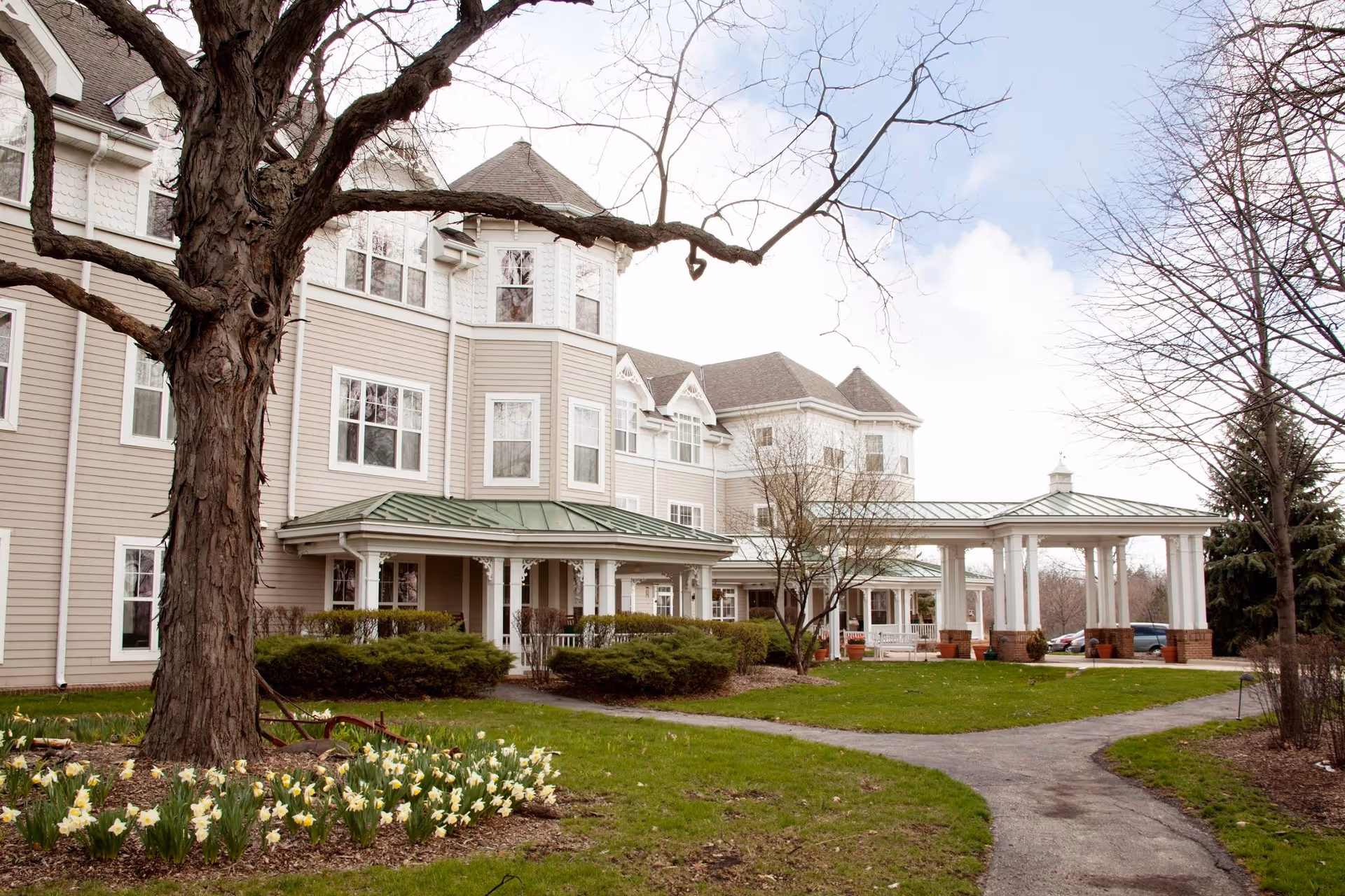 Front exterior of a multi-story senior living building with a covered entrance, landscaped lawn, and a large tree in the foreground.