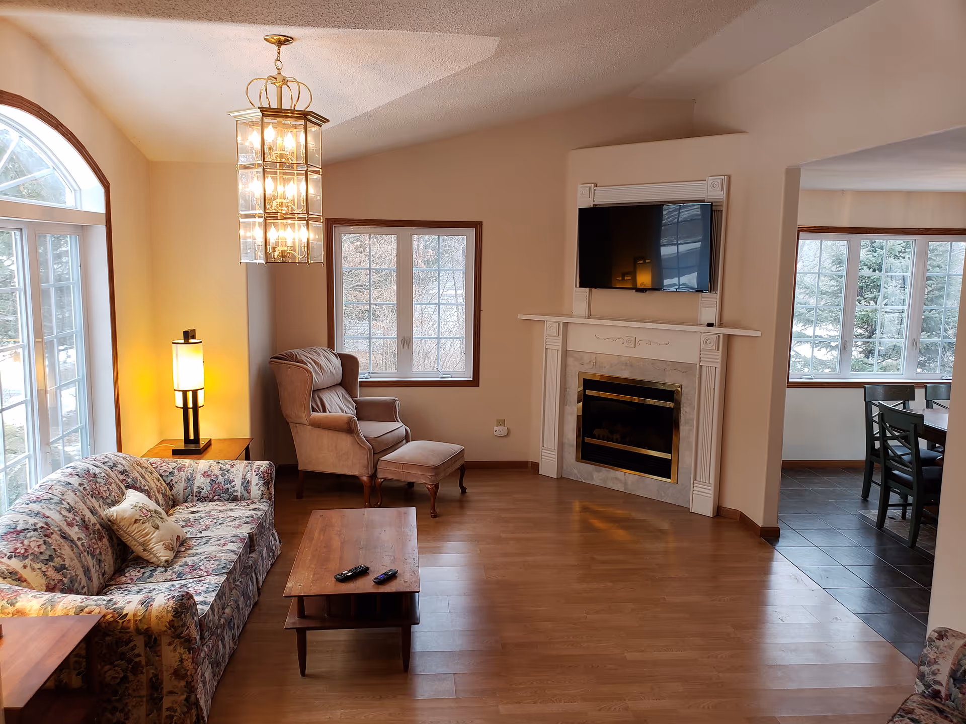 Bright living room with floral sofa, armchair, fireplace, TV, and large windows.