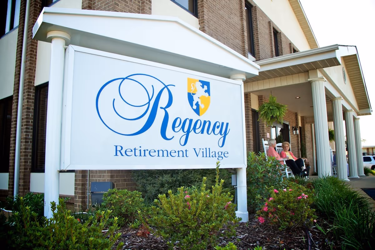 Exterior view of Regency Retirement Village building with a large white sign displaying the facility name. Two elderly women are sitting and talking on rocking chairs on the covered porch. There are green plants and flowers in the foreground.