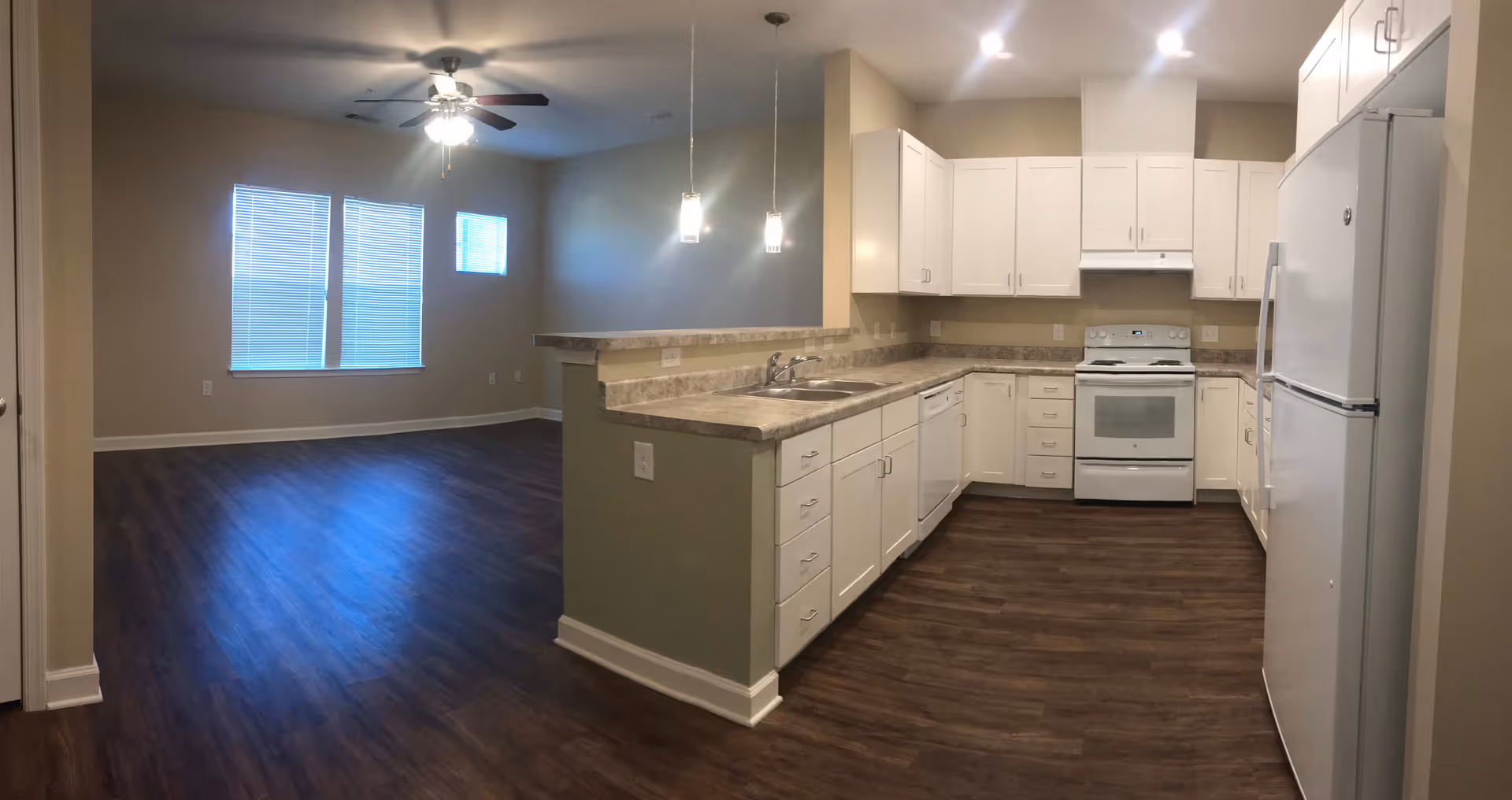 Open-concept interior showing a white kitchen with appliances and countertops opening to a living area with wood floors and windows.
