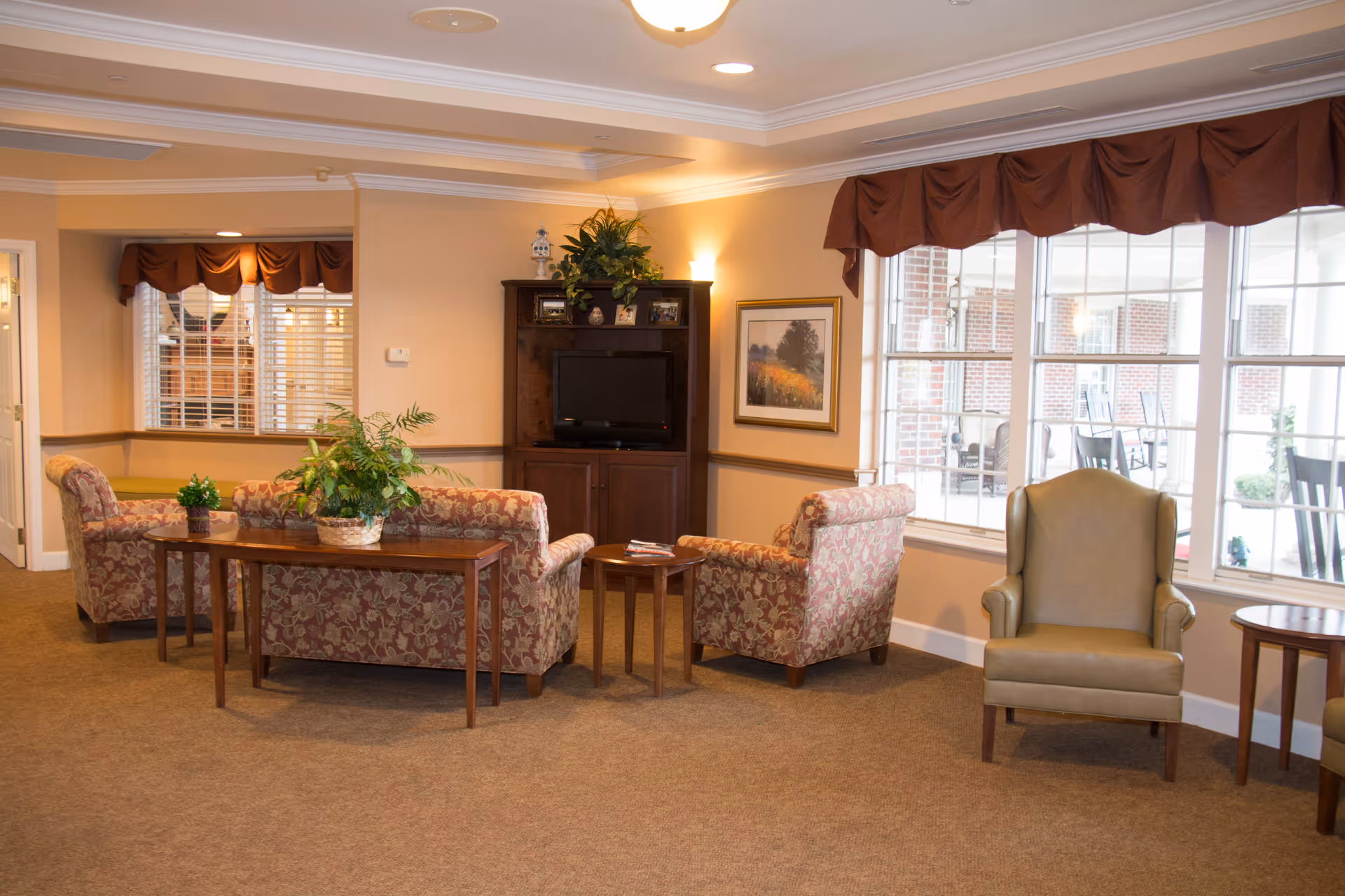 Cozy senior living lounge with floral sofas, tables, and a TV in a common area.