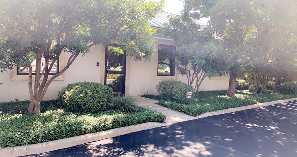 Single-story facility exterior with a sidewalk, entry door, and trimmed shrubs beneath trees.