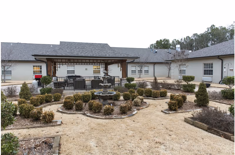 Outdoor courtyard area at a nursing and rehabilitation center featuring a central multi-tiered water fountain surrounded by neatly trimmed bushes and landscaped garden beds. In the background, there is a covered patio with outdoor seating and a barbecue grill, adjacent to a single-story building with multiple windows.