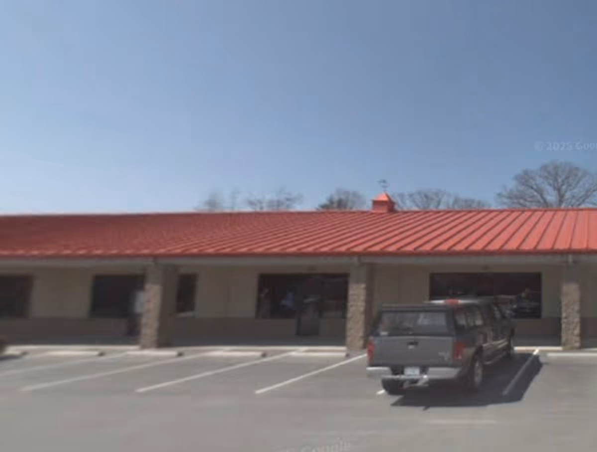 Front exterior of a single-story building with a red metal roof and parked pickup.