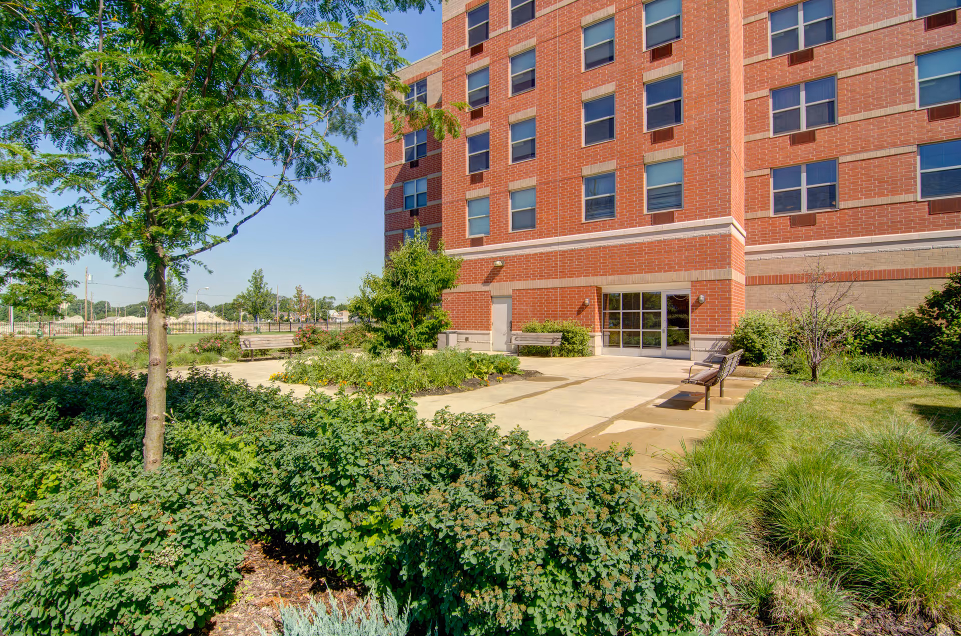 Exterior view of a red-brick senior living building with a landscaped courtyard, benches, and an entrance.