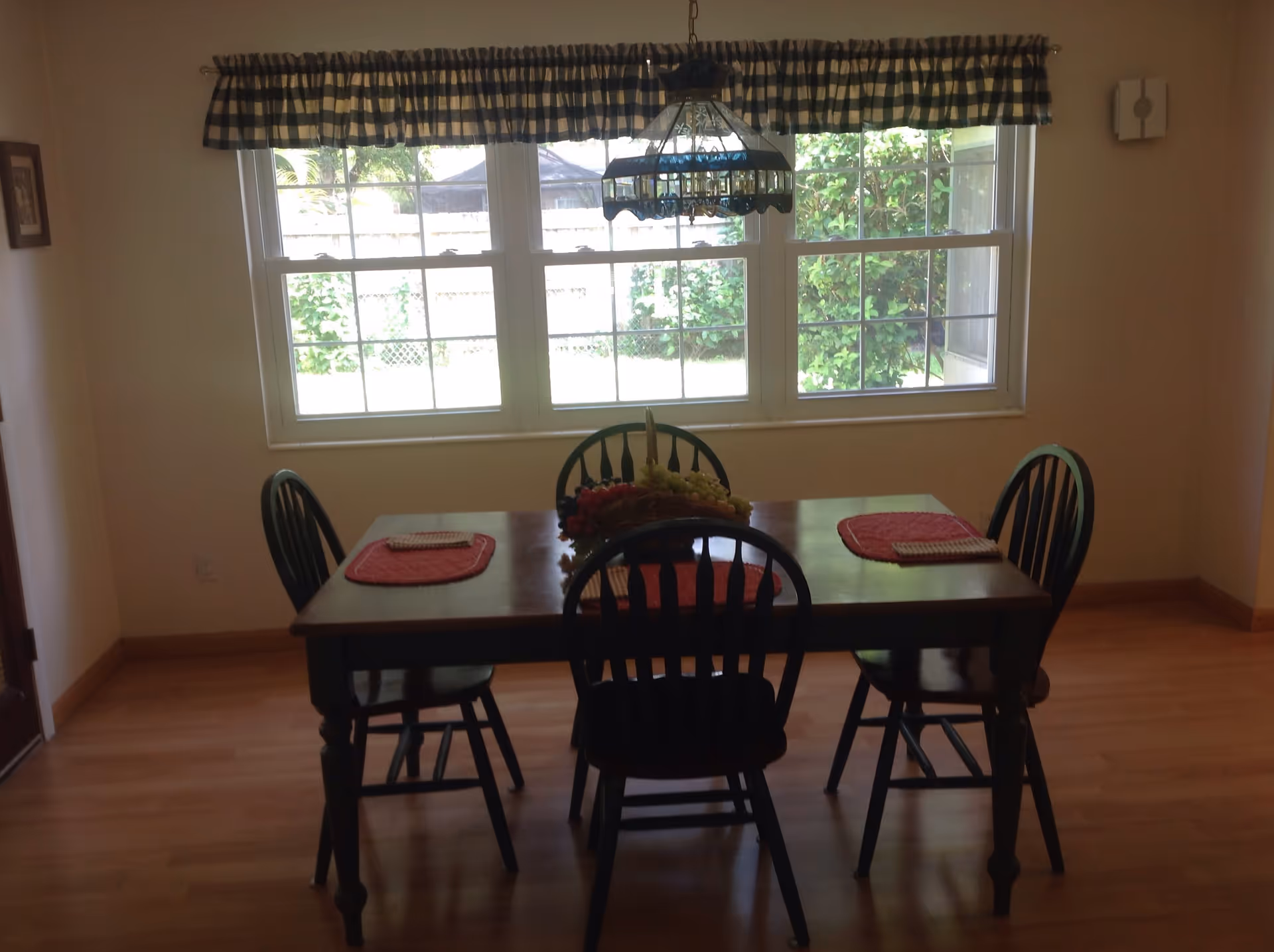 Wooden dining table with four chairs and placemats set beneath a hanging light in front of a large window with a blue-checked valance.