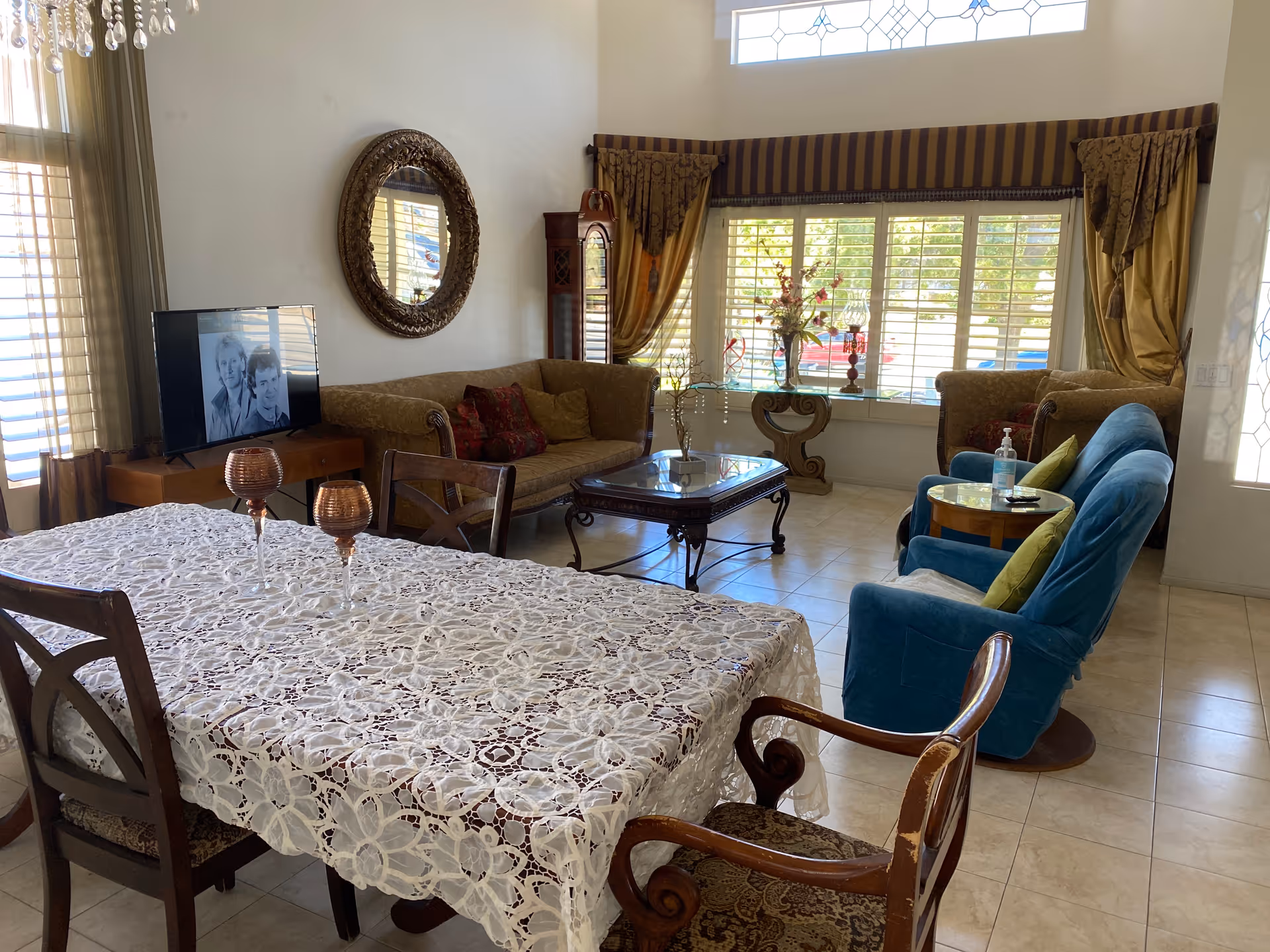A cozy living and dining area with a dining table covered by a white lace tablecloth and four wooden chairs. The living area has a brown sofa, a blue armchair with green pillows, a glass coffee table, and a TV on a wooden stand. There is a large window with striped curtains and a decorative round mirror on the wall.