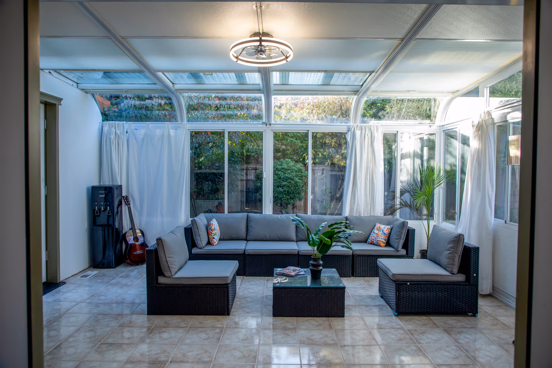 Bright glass-enclosed living room with gray cushioned wicker sectional and plants