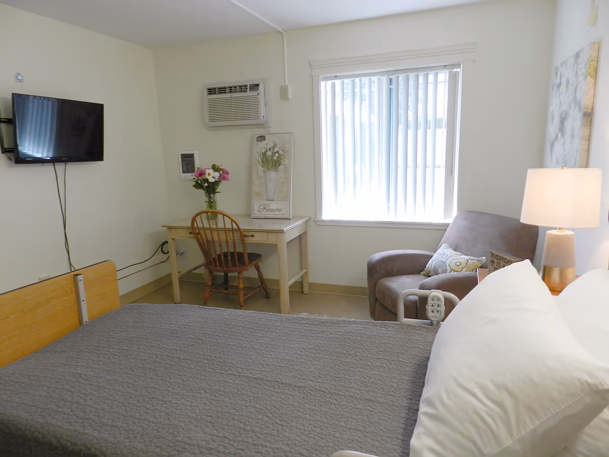 Simple patient bedroom with a bed in the foreground, a desk and chair, wall-mounted TV, armchair, and a window with vertical blinds.