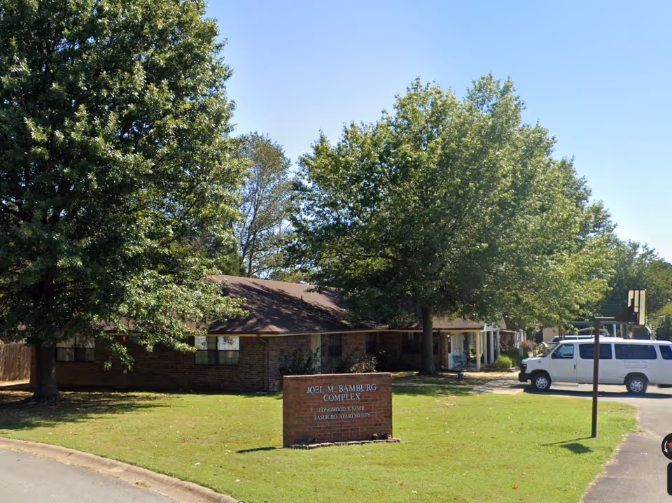 Brick senior living building with a sign, trees, and a white van outside.