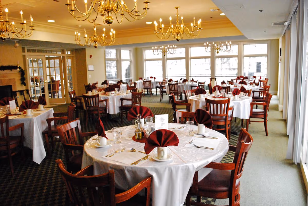 Bright dining room with round tables set with white tablecloths and folded red napkins under chandeliers.