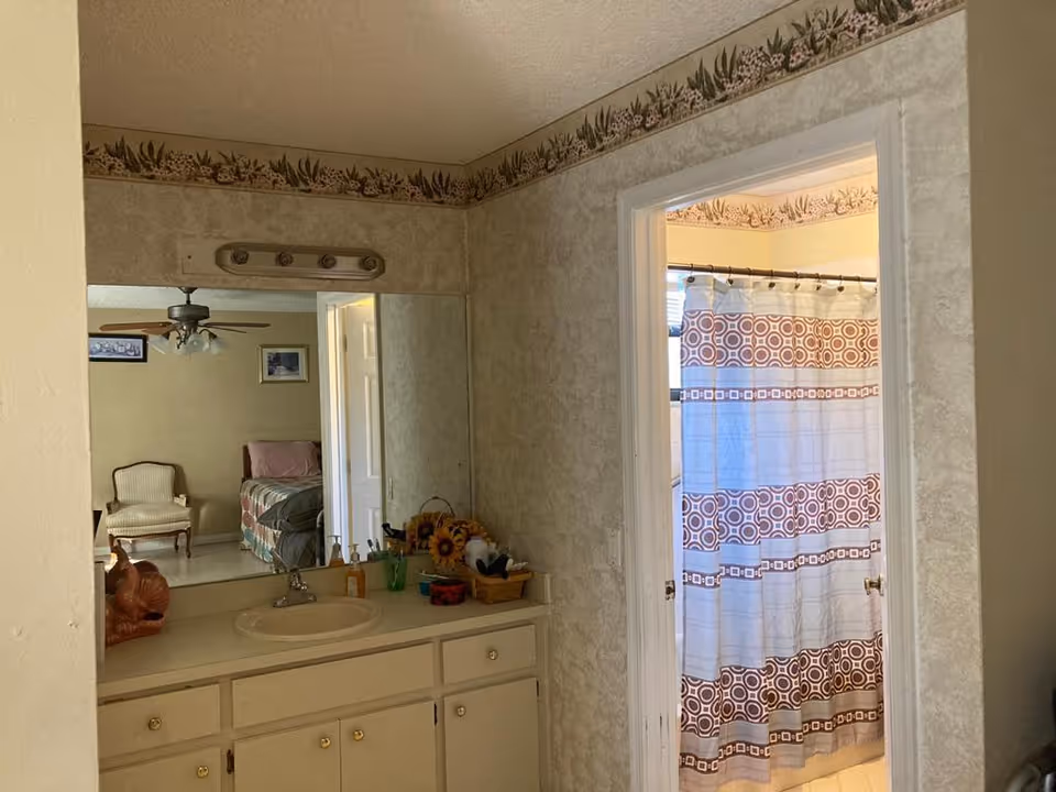 A bathroom vanity with a sink, mirror, and various toiletries on the counter. To the right, there is an open doorway leading to a bathroom with a shower curtain featuring a geometric pattern. The mirror reflects part of a bedroom with a chair, bed, ceiling fan, and framed pictures on the wall.