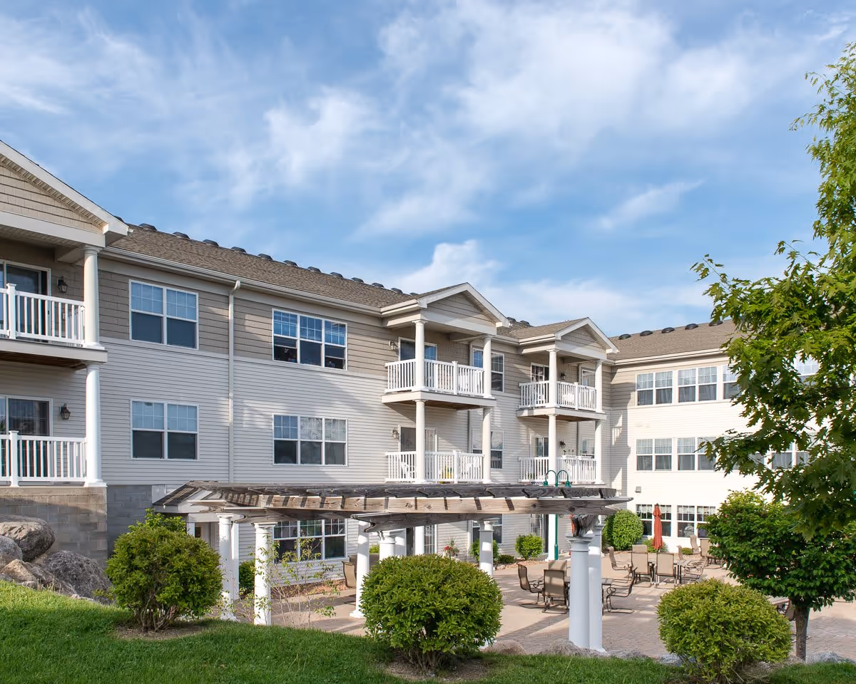 Exterior view of a three-story residential building with balconies, a pergola-covered patio and outdoor seating in a landscaped courtyard.