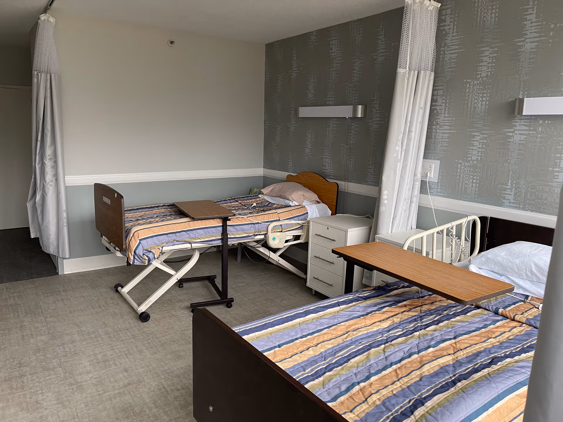 Two hospital-style beds in a rehab room with privacy curtains, a bedside table, and a dresser.