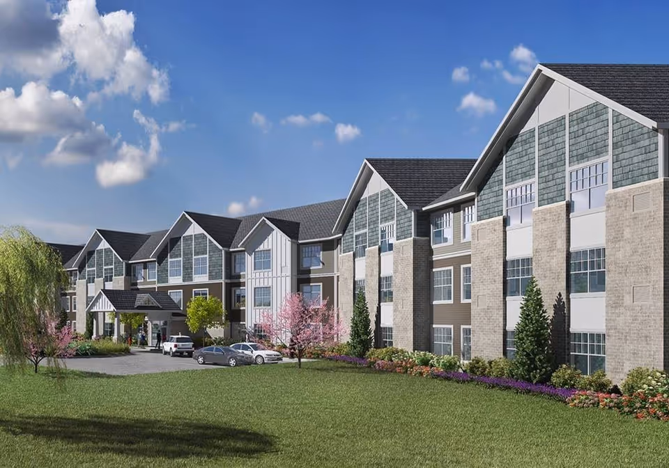 Exterior view of a large, modern senior living facility building with multiple windows, a covered entrance, parked cars, and landscaped greenery including trees and flower beds under a blue sky with some clouds.