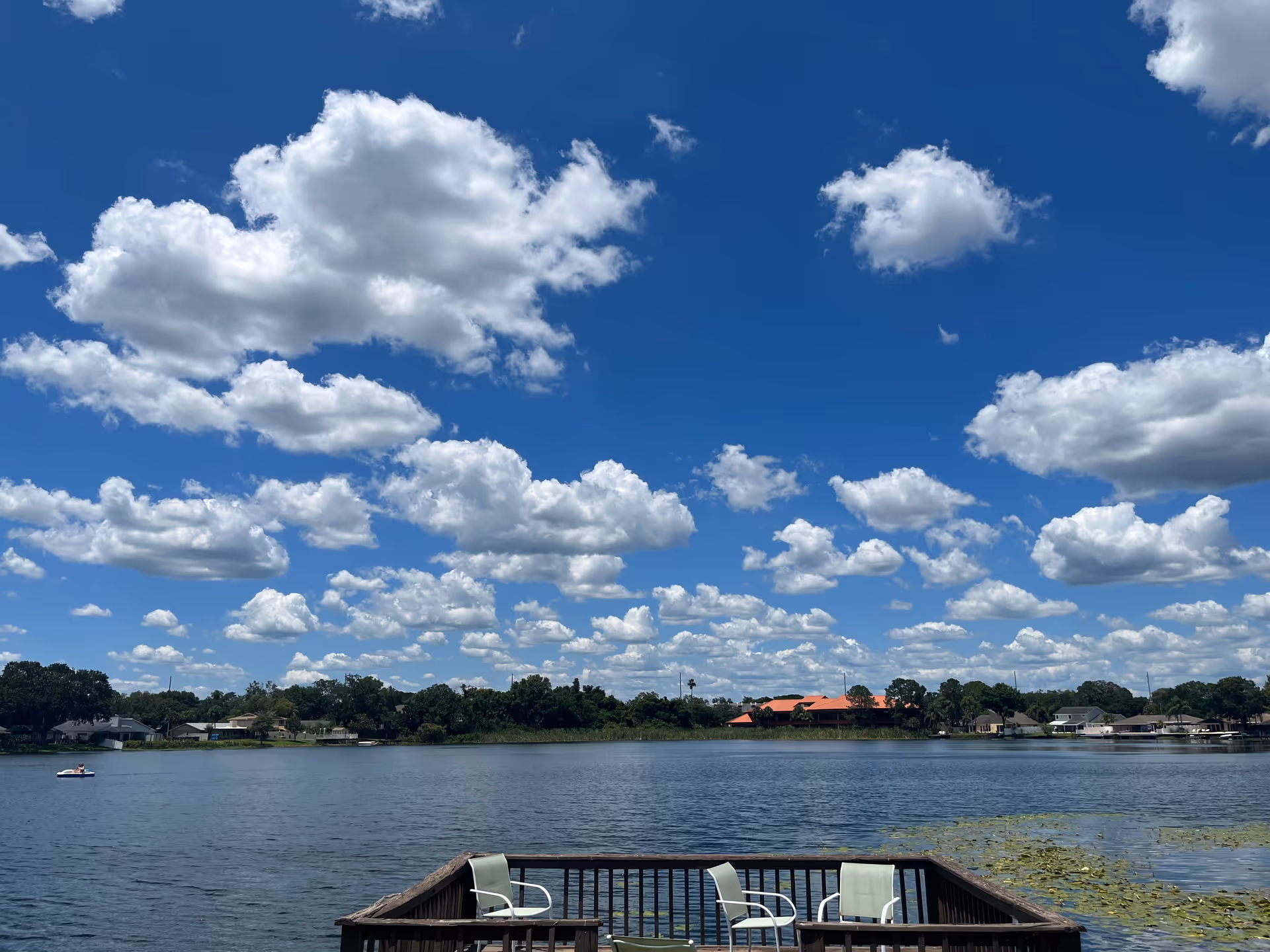 A scenic view of a lake under a bright blue sky filled with fluffy white clouds. In the foreground, there is a wooden deck with three light green chairs facing the water. Across the lake, there are houses and trees along the shoreline.