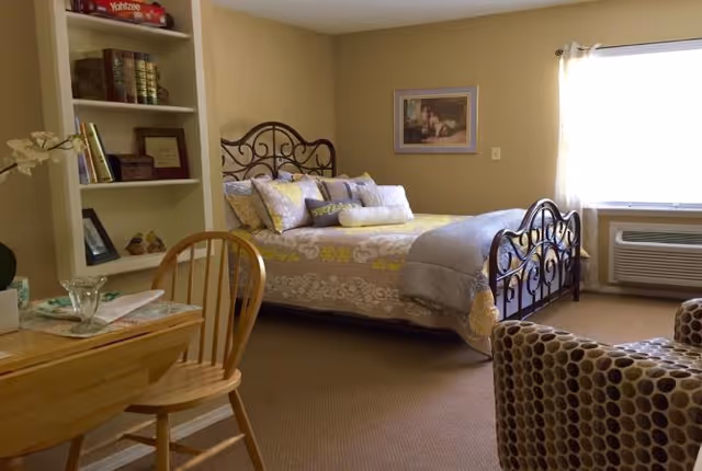 A cozy bedroom with a decorative metal bed frame, a bed made with patterned bedding and multiple pillows. There is a wooden chair and small table with glassware in the foreground, a built-in bookshelf with books and decor on the left, a framed picture on the wall, and a window with sheer curtains letting in natural light. An air conditioning unit is installed below the window.