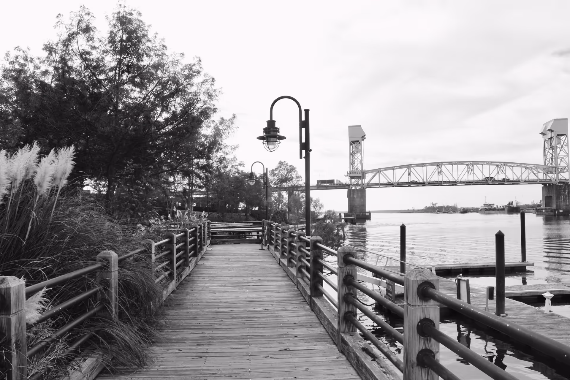 A wooden boardwalk along a riverfront with lamps, railing, trees, and a distant bridge