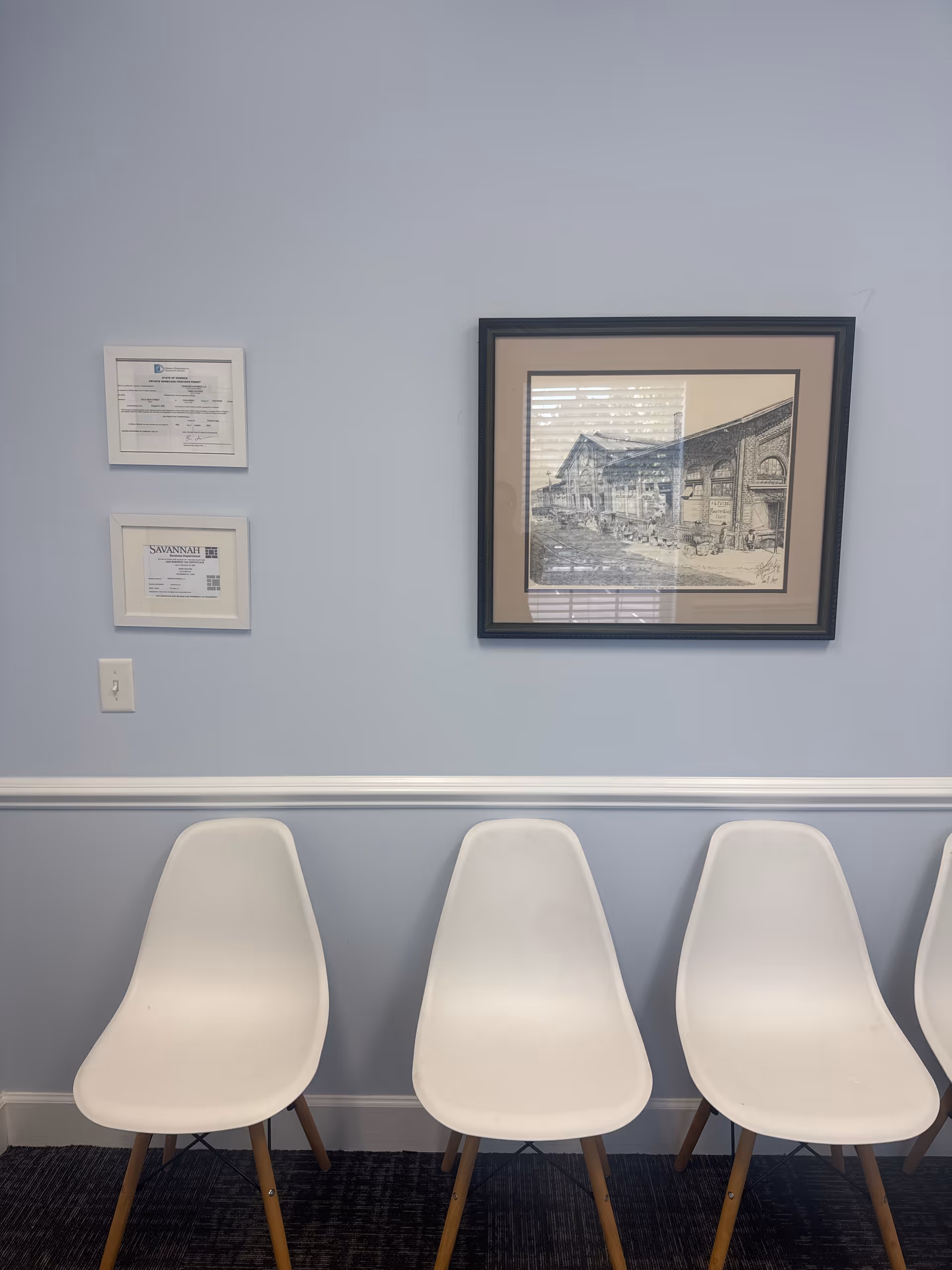 Blue-walled interior waiting area with white chairs lined along baseboard and framed artwork.
