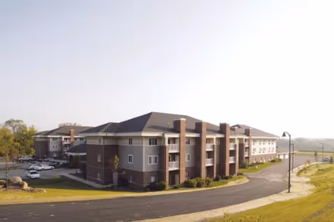 Exterior view of a large, multi-story senior living facility building with brick and gray siding, surrounded by a paved driveway and green lawn under a clear sky.