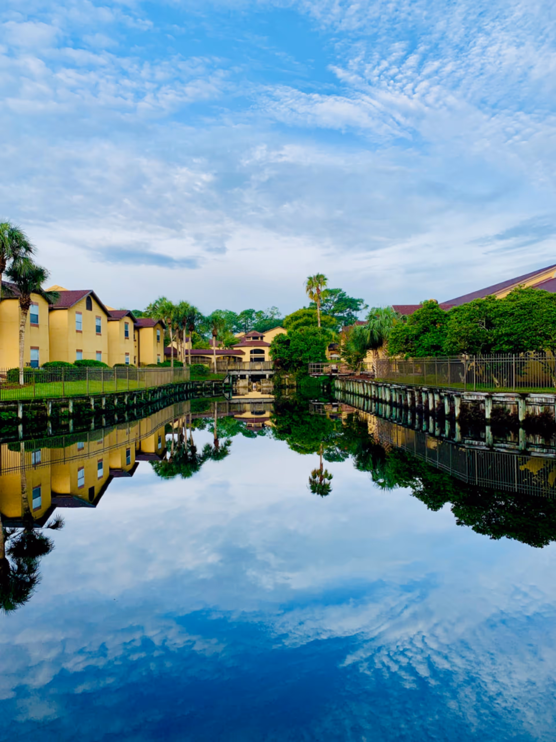 A serene outdoor scene at Fountains Rehabilitation at Mill Cove showing a calm water canal reflecting the blue sky, clouds, palm trees, and yellow buildings with red roofs on either side.