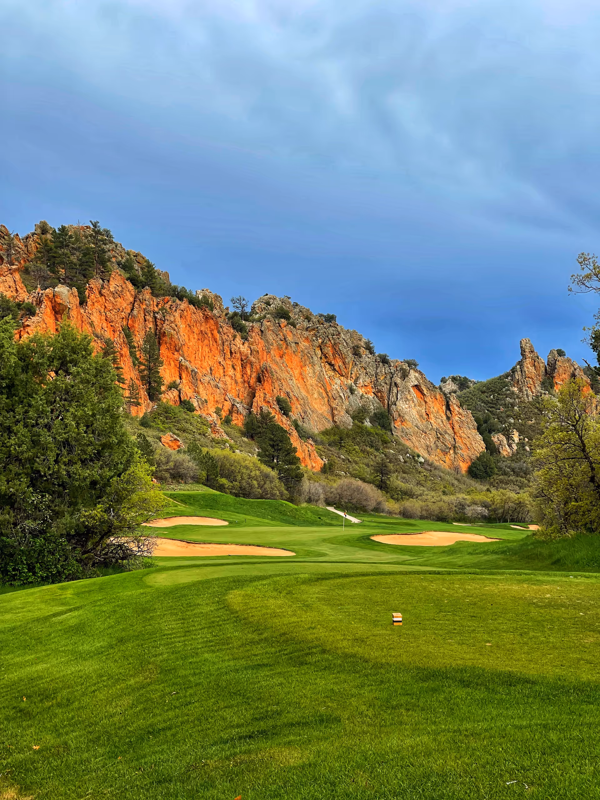 Lush green golf course winding past orange rocky cliffs beneath a cloudy blue sky.