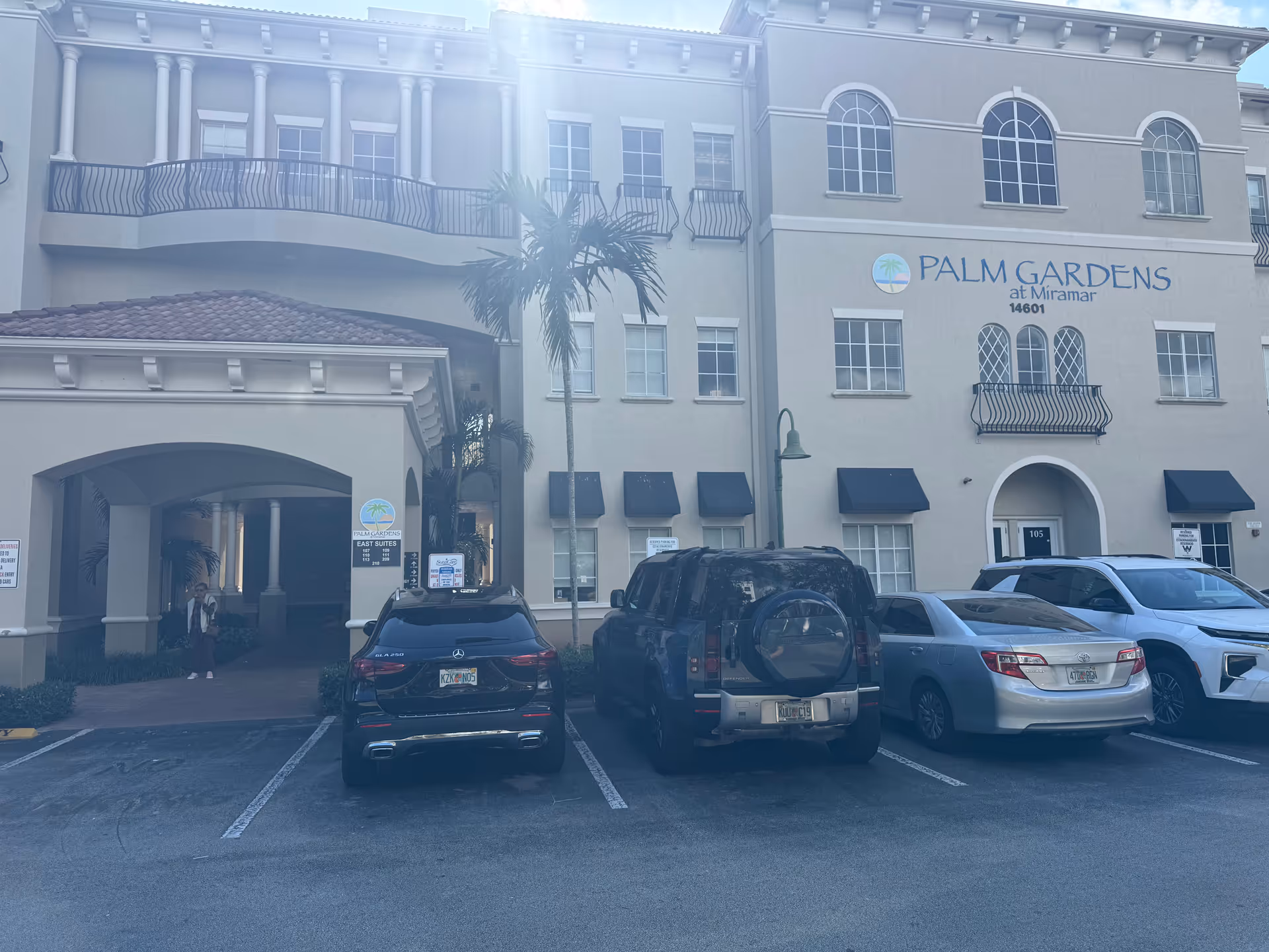 Exterior of Palm Gardens at Miramar, beige building with palm trees and parked cars.