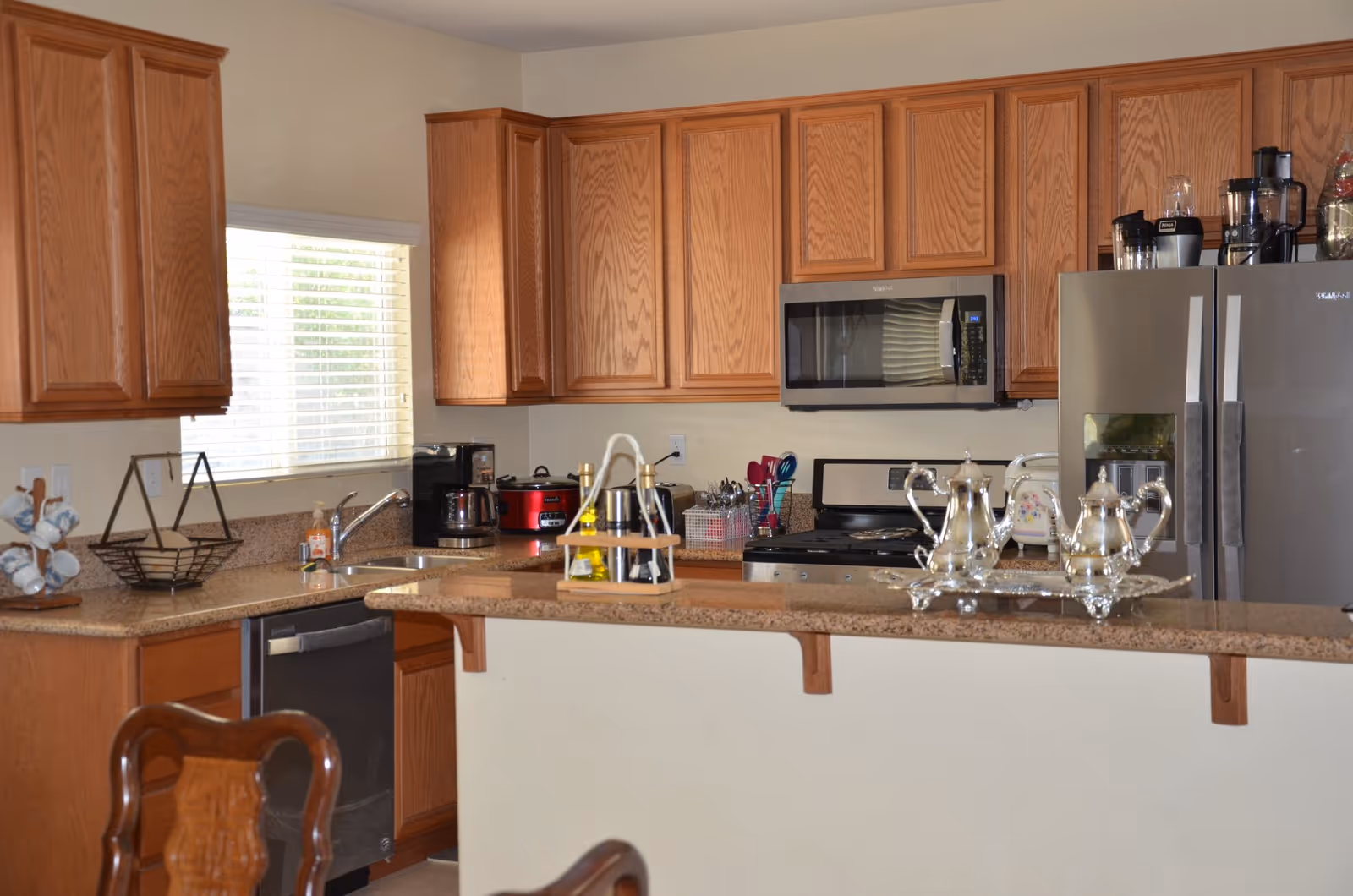 A kitchen with wooden cabinets, a granite countertop, and stainless steel appliances including a refrigerator, microwave, and dishwasher. The countertop has various items including a silver tea set, a spice rack, and kitchen utensils. A window with blinds lets in natural light.